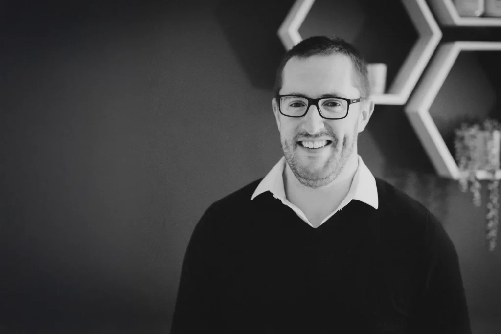 A black and white headshot of a man with short hair and a short beard, smiling warmly at the camera while wearing rectangular dark-framed glasses. He wears a dark jumper over a white collared shirt. Behind him, a wall-mounted hexagonal shelving unit is visible alongside a trailing plant, set against a dark background.
