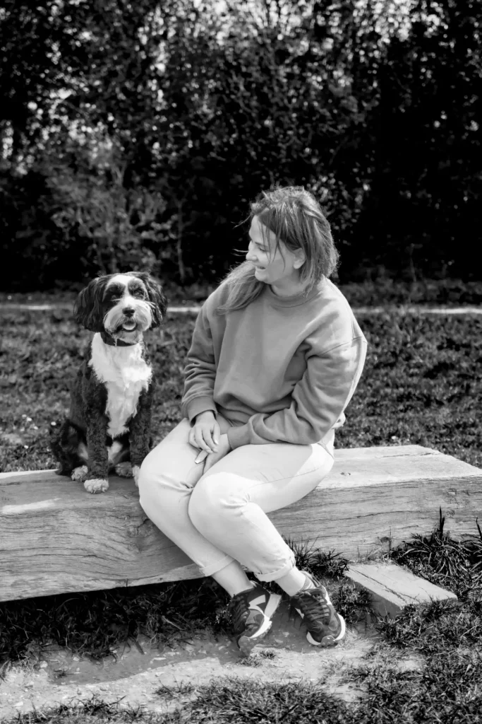 A black and white photograph of a woman with shoulder-length hair sitting on a large wooden log, smiling as she looks down at a black and white shaggy-coated dog seated beside her. She wears a crew-neck sweatshirt, cropped trousers, and trainers. The setting is outdoors on grass, with dense trees and shrubbery forming the backdrop.